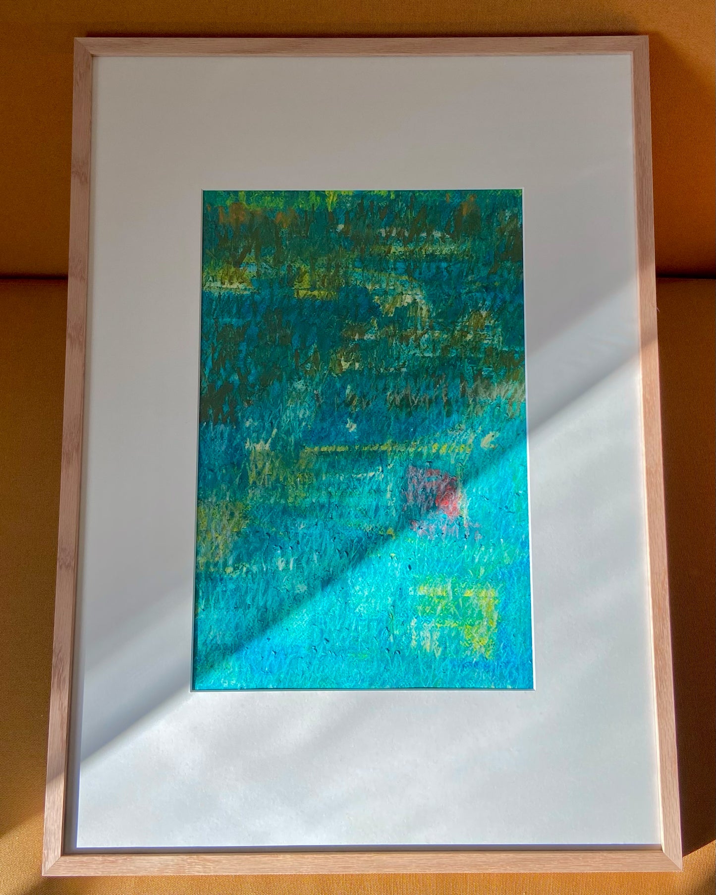 Mehrfarbige Chirografie in Ölpastell und Kreide auf Karton, gerahmt mit Passepartout und Holzrahmen, lehnend auf gelbem Polster, partiell mit Sonnenlicht beleuchtet
