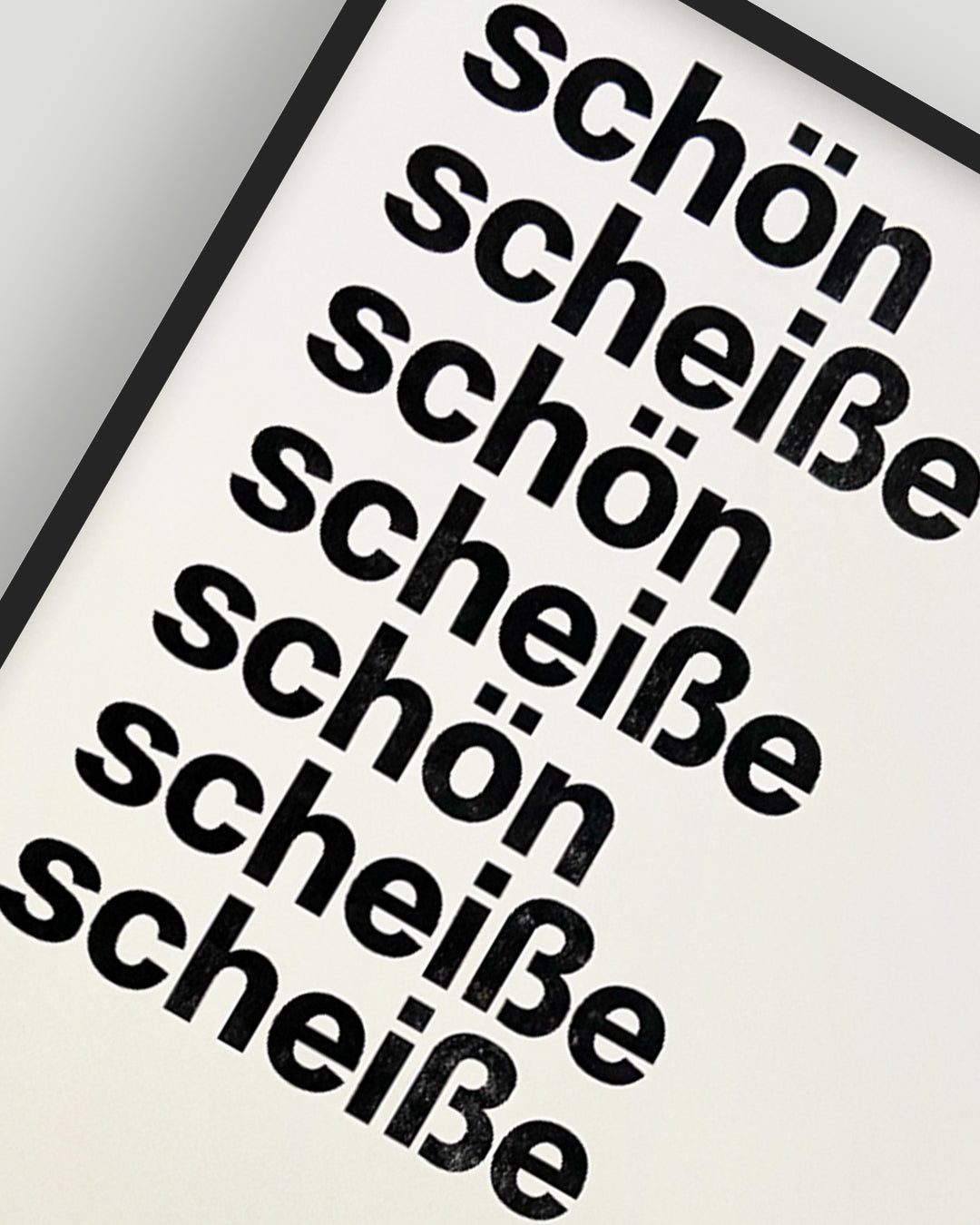 Nahaufnahme eines RISO-Druckes mit den Worten »schön« und »scheiße« in schwarzer Groteskschrift auf cremefarbenen Papier, gerahmt mit schwarzem Rahmen vor hellgrauem Hintergrund, nummeriert und signiert