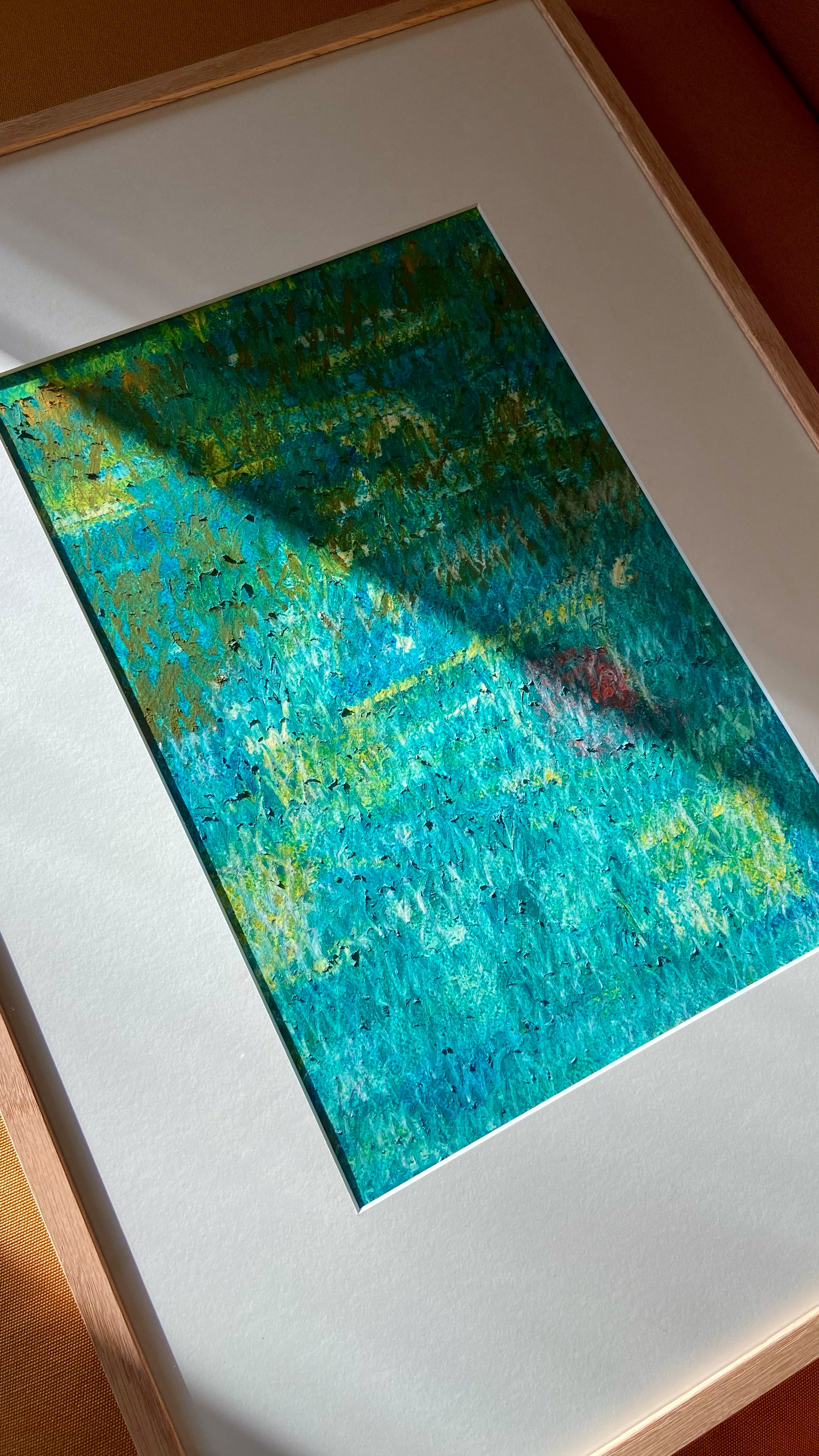 Mehrfarbige Chirografie in Ölpastell und Kreide auf Karton, gerahmt mit Passepartout und Holzrahmen, liegend auf gelbem Polster, partiell mit Sonnenlicht beleuchtet