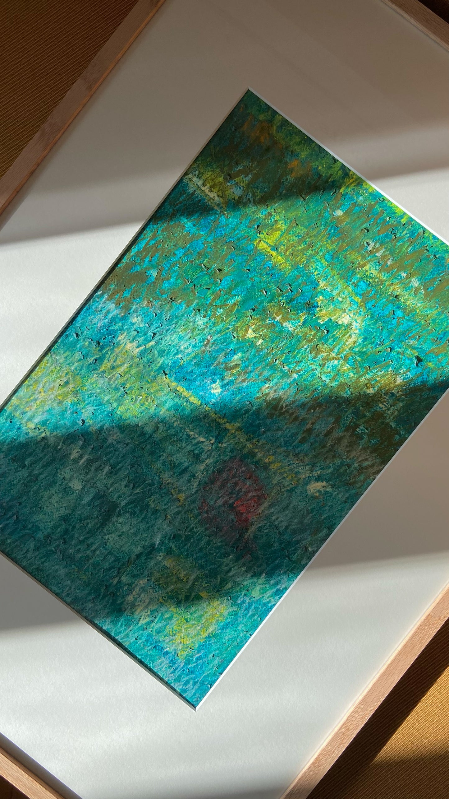 Mehrfarbige Chirografie in Ölpastell und Kreide auf Karton, gerahmt mit Passepartout und Holzrahmen, liegend auf gelbem Polster, partiell mit Sonnenlicht beleuchtet