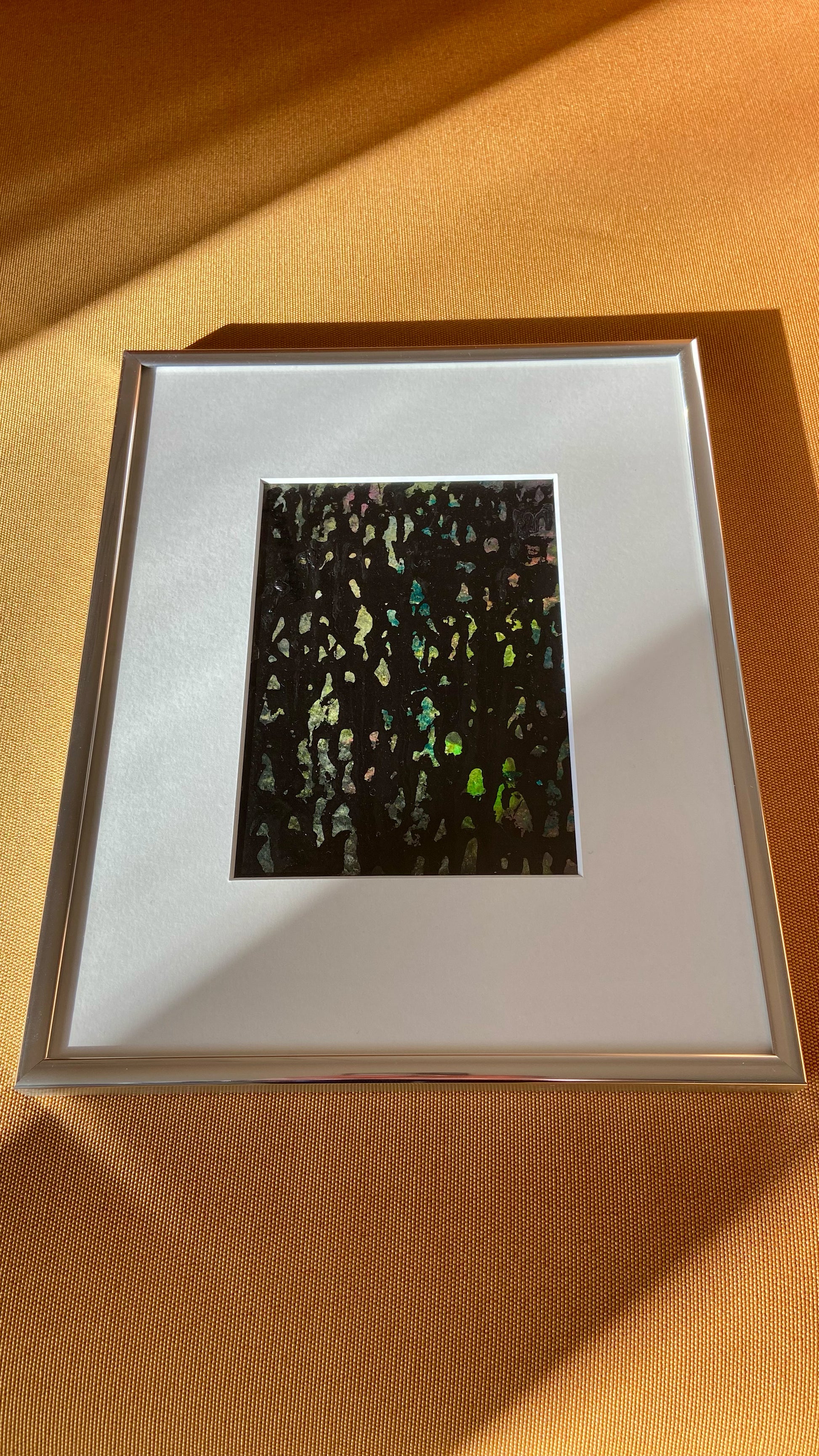 Mehrfarbige Chirografie in Acryl und Kreide auf Karton, gerahmt mit Passepartout und Aluminiumrahmen, liegend auf gelbem Polster, partiell mit Sonnenlicht beleuchtet