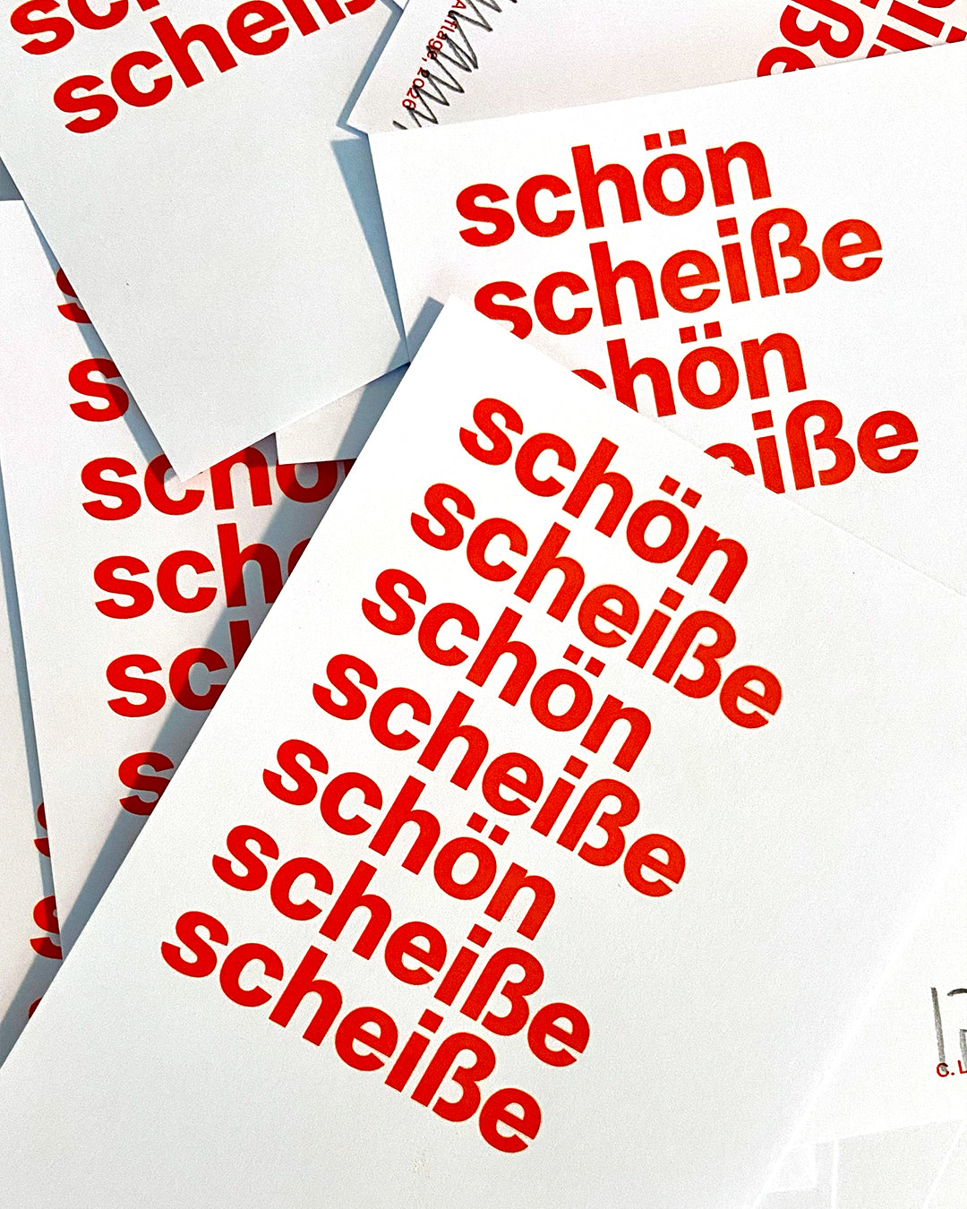 Mehrere Exemplare des roten Siebdrucks »schön scheiße« liegen überlappend auf einer Fläche und zeigen die typografische Gestaltung aus verschiedenen Blickwinkeln.