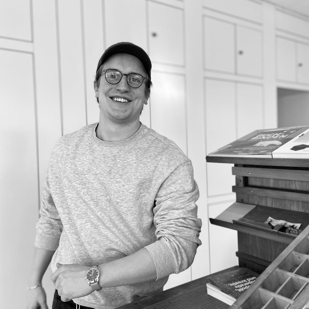 Portrait von Christoph Lehmann in einer Druckwerkstatt. Er trägt eine Kappe und Brille und lächelt in die Kamera.