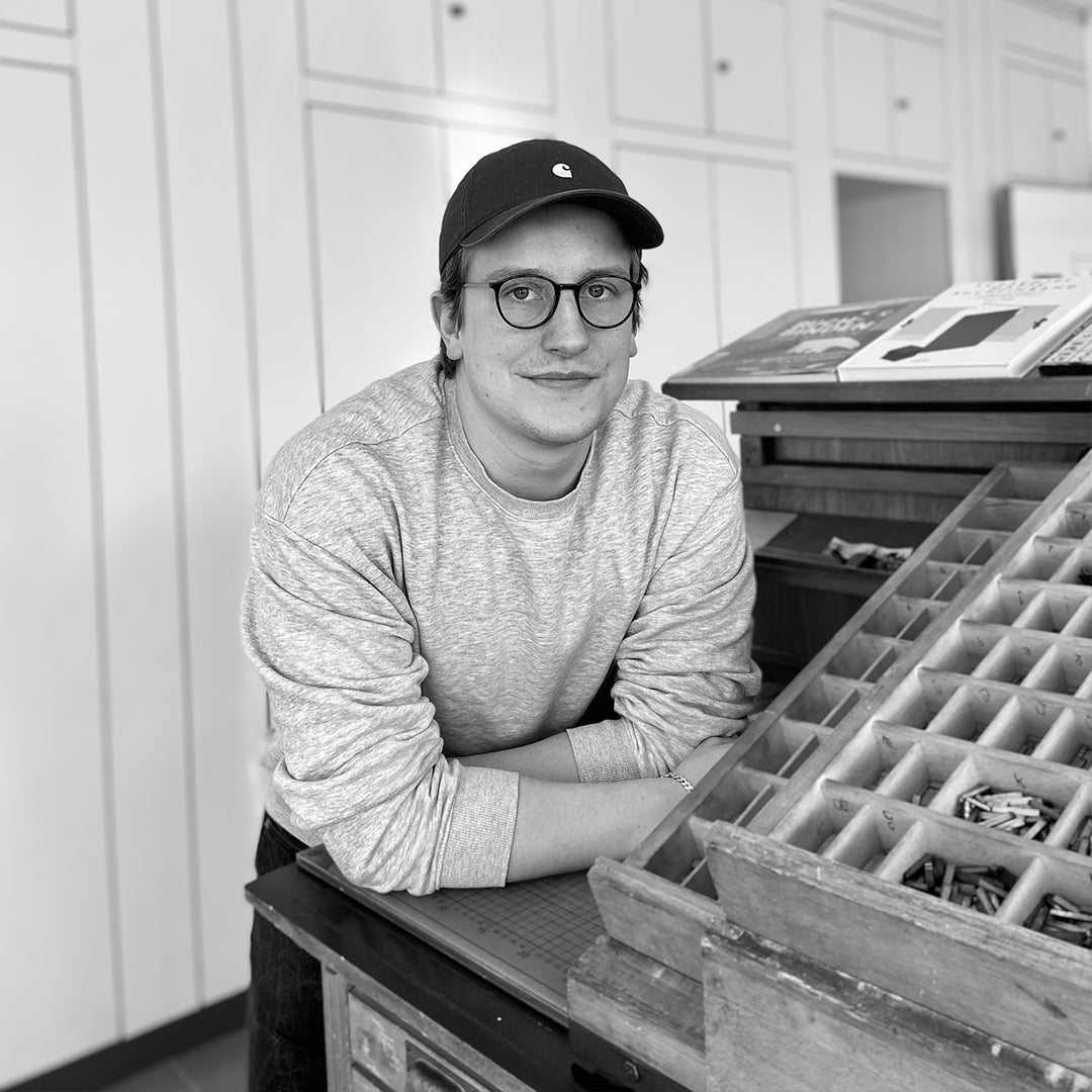 Portrait von Christoph Lehmann an einem Setzkasten in einer Bleisatzwerkstatt. Er trägt eine Kappe und Brille und blickt in die Kamera.